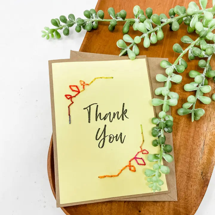 Yellow card with &#39;Thank You&#39; text and embroidered leaves on a wooden surface with greenery.